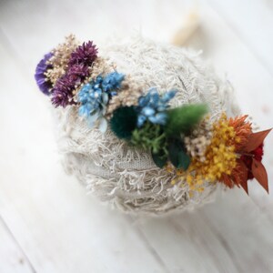 Puede incluir: Una diadema de crochet blanco adornada con un arco&iacute;ris de flores secas. Las flores son moradas, azules, verdes, amarillas, naranjas y rojas.