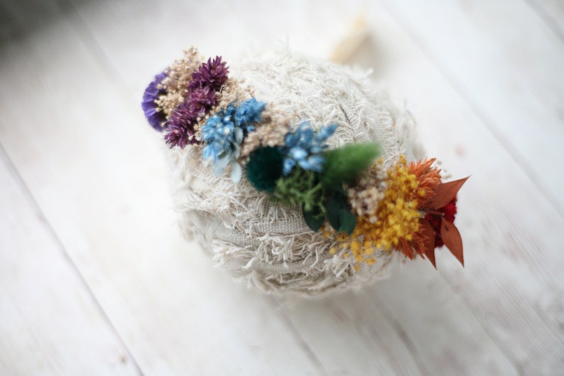 Puede incluir: Una diadema de crochet blanco adornada con un arco&iacute;ris de flores secas. Las flores son moradas, azules, verdes, amarillas, naranjas y rojas.