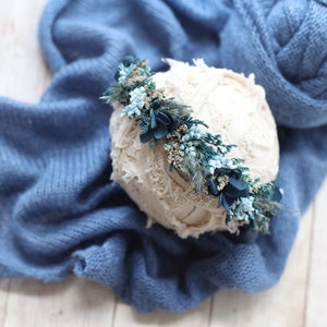 May include: A cream-colored, textured prop for newborn photography, adorned with a floral crown of teal and blue flowers. The prop rests on a soft, blue knitted blanket. The scene is set on a light wood surface, creating a gentle, natural aesthetic.