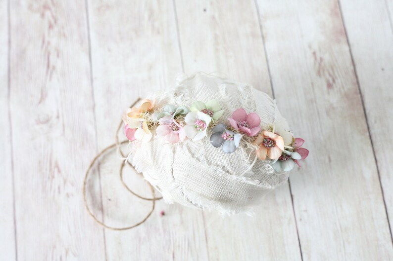 Puede incluir: Una corona floral con flores de tela en tonos pastel, rosa, melocot&oacute;n y azul. Las flores est&aacute;n dispuestas sobre una base blanca texturizada, con un acento de cuerda r&uacute;stica. La corona es para un beb&eacute; o ni&ntilde;o.