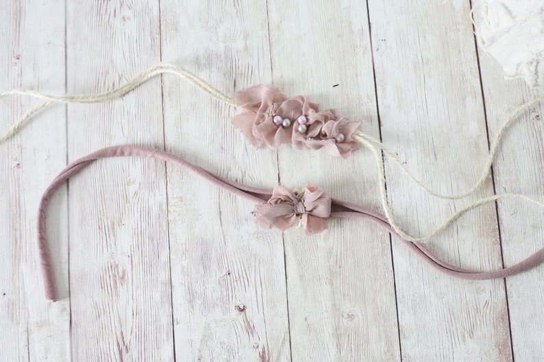 May include: Two baby headbands. One is a dusty rose color with a ruffled fabric design and small pearls. The other is a dusty rose headband with a small bow. Both headbands have a soft, stretchy fabric.
