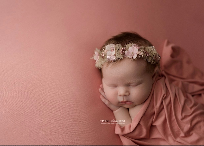 May include: A newborn baby girl sleeping peacefully on a pink blanket, wearing a flower crown with pink and white flowers.