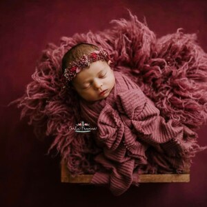May include: A newborn baby girl is sleeping peacefully in a pink fuzzy blanket. She is wearing a pink headband with flowers. The background is a deep burgundy color.