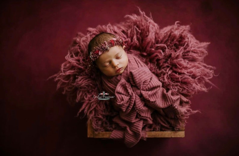 May include: A newborn baby girl is sleeping peacefully in a pink fuzzy blanket. She is wearing a pink headband with flowers. The background is a deep burgundy color.