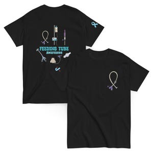 以下が含まれることがあります： 栄養チューブの啓発に関連するデザインの黒いTシャツ。1枚のシャツには、医療機器のイラストとともに「FEEDING TUBE Awareness」の文字がプリントされています。もう1枚のシャツには、栄養チューブの小さなデザインがあります。