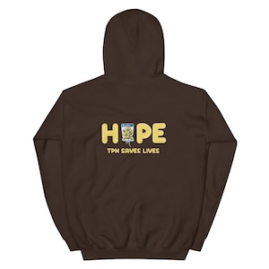 Può includere: Felpa con cappuccio marrone scuro. Sul retro, la parola "HOPE" in lettere gialle, con un'immagine floreale e la scritta "TPN SAVES LIVES" sotto. Un capo casual e comodo.