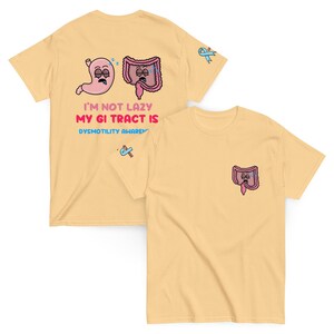 May include: Two light yellow t-shirts. One shirt has a graphic of a sleeping stomach and colon with the text "I'M NOT LAZY MY GI TRACT IS DYSMOTILITY AWARENESS". The other shirt has a pocket with a colon graphic.