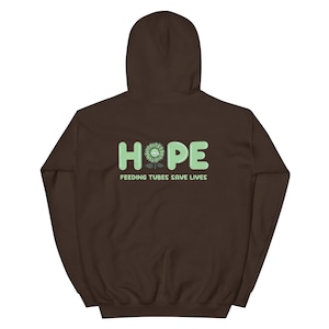 Könnte beinhalten: Ein brauner Kapuzenpullover mit dem Wort "HOPE" in hellgrünen Blockbuchstaben. Eine Sonnenblume ersetzt das "O". Unter dem Wort "HOPE" steht der Text "FEEDING TUBES SAVE LIVES" in Hellgrün. Der Kapuzenpullover hat eine Kapuze und lange Ärmel.