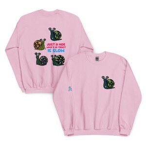 May include: Pink sweatshirt featuring a cartoon snail with a green and yellow shell. The text "JUST A HOE WHO'S GI TRACT IS SLOW" is printed on the back. A smaller snail graphic is on the front.