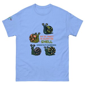 May include: Light blue t-shirt featuring four cartoon snails with various shell patterns. The text "MY GI TRACT IS SLOW AS SHELL" and "DYSMOTILITY AWARENESS" are printed on the shirt. The shirt has short sleeves and a crew neck.