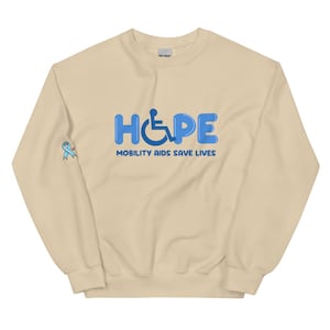 Könnte beinhalten: Beiges Sweatshirt mit dem Wort "HOPE" in blauen Buchstaben mit Blasen-Effekt, mit einem Rollstuhlsymbol. Darunter steht der Text "MOBILITY AIDS SAVE LIVES". Auf dem linken Ärmel befindet sich ein hellblaues Band. Dieses Sweatshirt fördert das Bewusstsein für Behinderungen.