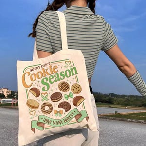 May include: Cream-colored tote bag with the text "Sorry Can't Cookie Season" and illustrations of various cookies. The bag has a green and orange color scheme, with a light green ribbon at the bottom that reads "How Many Boxes?" The bag has a cream-colored strap.