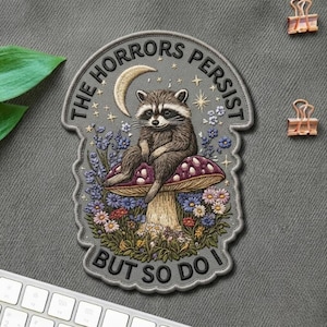 May include: Embroidered patch featuring a raccoon seated on a mushroom, surrounded by flowers and the text "THE HORRORS PERSIST BUT SO DO I." The patch has a gray background with a crescent moon and stars.