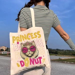 May include: Cream-colored tote bag with a graphic of a cat wearing sunglasses and a crown, with the words "PRINCESS" and "DONUT" in yellow and pink. The bag has a simple design with a rectangular shape and two handles.