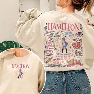 May include: Cream-colored sweatshirt featuring the word "HAMILTON" and a graphic of a figure on a star. The back of the sweatshirt has a larger design with musical notes, text, and illustrations related to the musical.