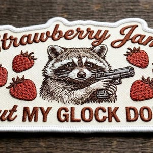 May include: Embroidered patch with a raccoon holding a gun, surrounded by strawberries. Text reads "Strawberry Jams But MY GLOCK DON'T" in brown thread on a white background.