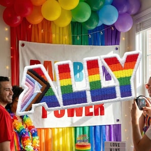 May include: A colorful embroidered "ALLY" sign with a rainbow design, displayed against a backdrop of rainbow-colored decorations, balloons, and a banner that reads "POWER". The scene is festive and celebratory.