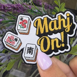 May include: Embroidered patch featuring the text "Mahj On!" in yellow and black, with three Mahjong tiles. The tiles display floral and Chinese character designs. The patch is white with a black border.