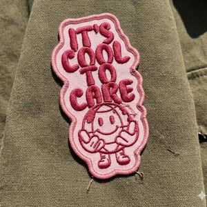 May include: A pink embroidered patch with the words "IT'S COOL TO CARE" in a bubble font. The patch features a smiling globe character with arms and legs. The patch is sewn onto an olive green fabric.