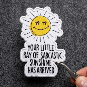 May include: Embroidered patch featuring a smiling yellow sun with white petals and the text "YOUR LITTLE RAY OF SARCASTIC SUNSHINE HAS ARRIVED" in black lettering. The patch is white with black and yellow accents.