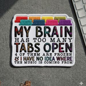 May include: Embroidered patch with the text "MY BRAIN HAS TOO MANY TABS OPEN." The patch has a white background with colorful tabs across the top. The text also reads "4 OF THEM ARE FROZEN & I HAVE NO IDEA WHERE THE MUSIC IS COMING FROM."