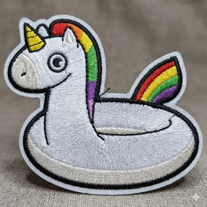 May include: Embroidered patch of a white unicorn floating on a pool float. The unicorn has a yellow horn, a rainbow mane, and a rainbow wing. The patch is outlined in black.