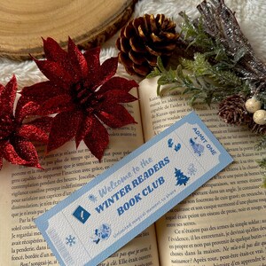 Può includere: Un segnalibro azzurro con la scritta "Welcome to the Winter Readers Book Club" e illustrazioni a tema invernale. Il segnalibro è posto su un libro aperto, con stelle di Natale rosse e una pigna sullo sfondo.