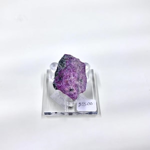 May include: A rough-cut, natural purple mineral specimen with dark gray and black inclusions. The stone is displayed on a clear acrylic stand against a white background. A small white tag with the price is visible.