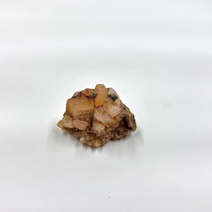 May include: A cluster of raw, unpolished crystals in shades of tan, peach, and brown. The crystals have a rough, textured surface and are clustered together on a darker brown base. The crystals are illuminated by natural light.