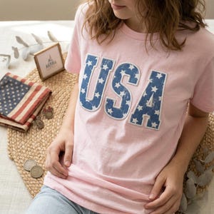 Könnte beinhalten: Hellrosa T-Shirt mit dem Wort "USA" in blauen Buchstaben, gefüllt mit weißen Sternen. Das Bild zeigt patriotische Accessoires, wie eine kleine amerikanische Flagge und dekorative Gegenstände.