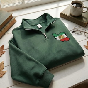 May include: A forest green sweatshirt with a quarter-zip neckline. The sweatshirt features a colorful embroidered patch of the Iranian flag on the chest. The patch is in the shape of the country. The sweatshirt is made of a soft material.