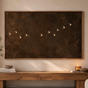 May include: A rectangular wall art piece featuring a dark brown, textured background with a flock of small, light-coloured birds flying in a diagonal formation across the centre, all enclosed within a thin, metallic-toned frame.