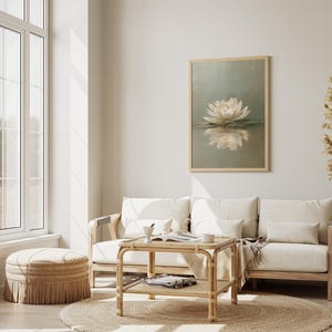 May include: A serene living room scene featuring a framed artwork of a white lotus flower reflected in water. The room includes a light-colored sofa, a woven coffee table, a round jute rug, and a decorative ottoman. Dried pampas grass adds a natural touch.