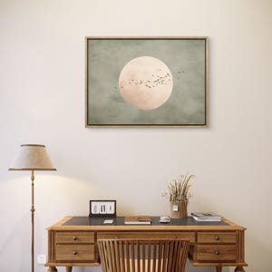 May include: A framed artwork featuring a large, pale pink circle against a muted green background, with a flock of birds in silhouette. The artwork hangs above a wooden desk with a matching chair, a lamp, and decorative items.
