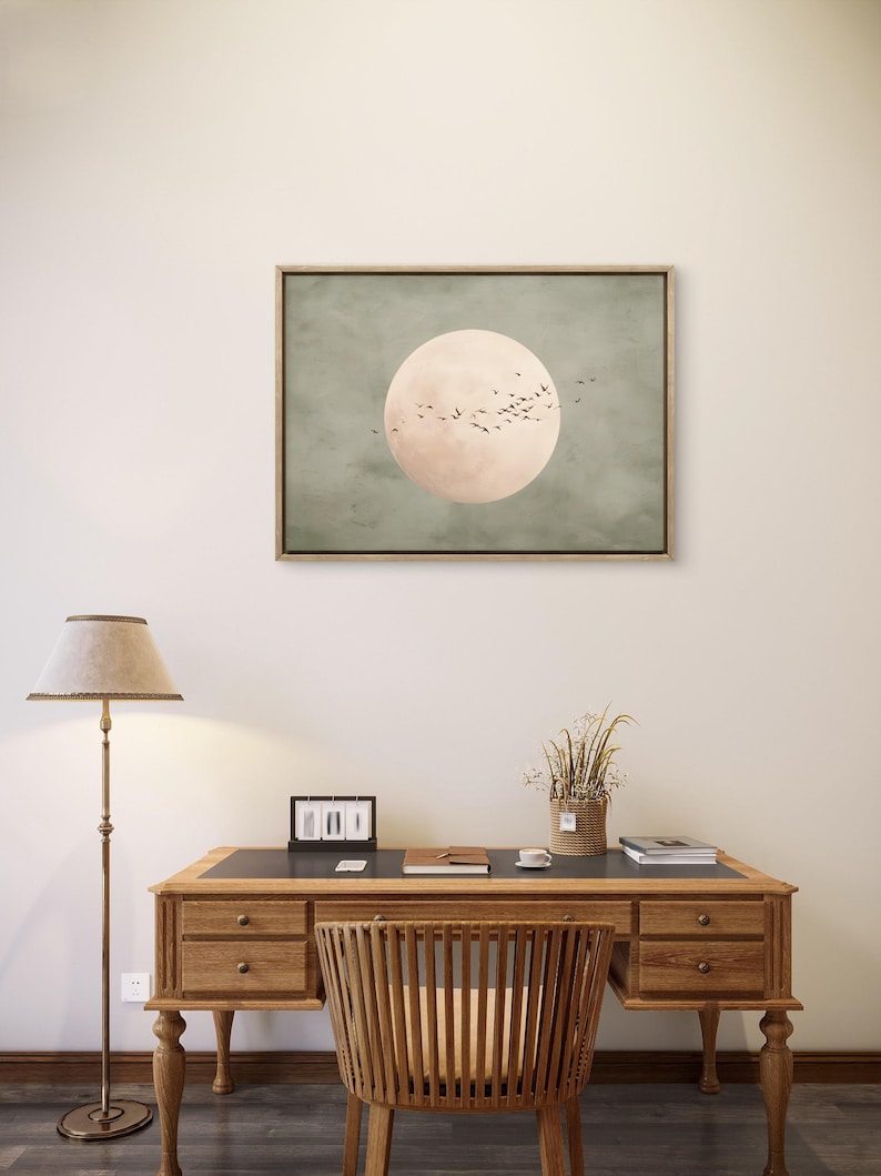 May include: A framed artwork featuring a large, pale pink circle against a muted green background, with a flock of birds in silhouette. The artwork hangs above a wooden desk with a matching chair, a lamp, and decorative items.