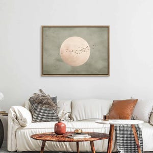 May include: A framed canvas art print featuring a large, pale peach-colored circle with a flock of birds in flight against a muted green and gray background. The artwork is displayed above a white sofa.