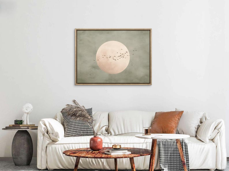 May include: A framed canvas art print featuring a large, pale peach-colored circle with a flock of birds in flight against a muted green and gray background. The artwork is displayed above a white sofa.