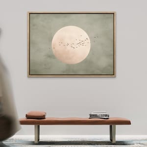 May include: A framed artwork depicts a large, pale peach-colored circle, resembling a moon, set against a muted green background. Silhouetted birds fly across the moon. A brown bench with a small pillow and books sits below the artwork.