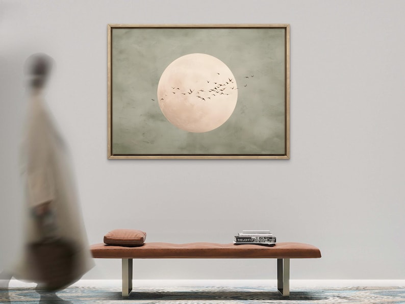 May include: A framed artwork depicts a large, pale peach-colored circle, resembling a moon, set against a muted green background. Silhouetted birds fly across the moon. A brown bench with a small pillow and books sits below the artwork.