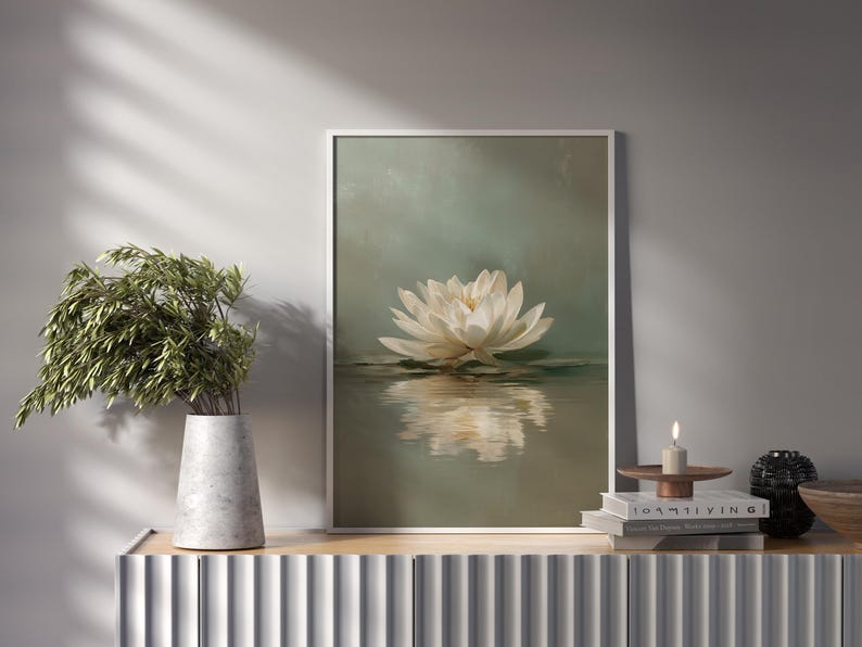 May include: A framed art print featuring a white lotus flower floating on water. The artwork has a soft, muted color palette with a reflective surface. A vase with greenery and a candle are on a nearby surface.