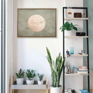 May include: A framed artwork depicts a large, pale pink circle with a flock of birds against a muted green background. The artwork hangs above a gray shelf holding three potted plants. A tall, black and white shelf displays various decorative items and plants.
