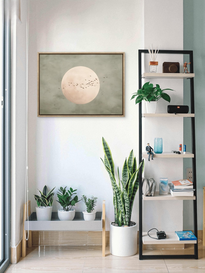 May include: A framed artwork depicts a large, pale pink circle with a flock of birds against a muted green background. The artwork hangs above a gray shelf holding three potted plants. A tall, black and white shelf displays various decorative items and plants.