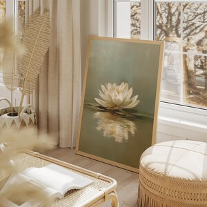 May include: A framed art print featuring a white water lily floating on water. The artwork has a soft, muted color palette with a light wooden frame. The scene is set in a bright, airy room with natural light.
