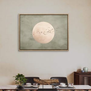 May include: A framed artwork featuring a large, pale pink circle resembling a moon against a muted green background. Silhouetted birds fly across the moon's surface. The artwork is displayed above a dining table set with dishes and a centerpiece.