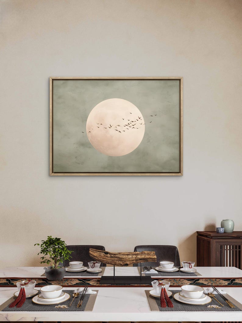 May include: A framed artwork featuring a large, pale pink circle resembling a moon against a muted green background. Silhouetted birds fly across the moon's surface. The artwork is displayed above a dining table set with dishes and a centerpiece.