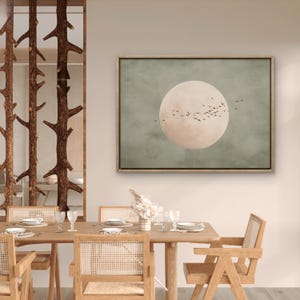 May include: A framed canvas art print featuring a large, pale pink moon against a muted green background. Silhouetted birds fly across the moon. The artwork hangs above a wooden dining table with woven chairs, creating a serene, nature-inspired aesthetic.