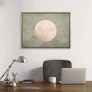 May include: A framed art print featuring a large, pale peach-colored circle, possibly representing the moon, set against a muted green background. Silhouettes of birds are in flight around the circle. The frame is a light wood color.