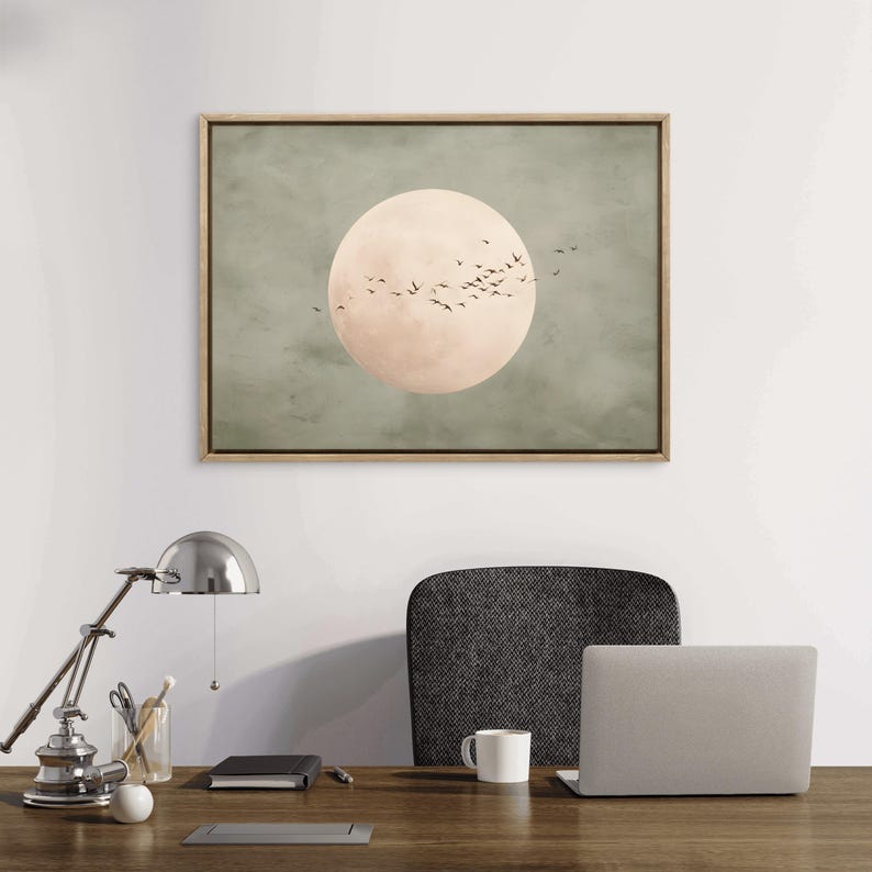 May include: A framed art print featuring a large, pale peach-colored circle, possibly representing the moon, set against a muted green background. Silhouettes of birds are in flight around the circle. The frame is a light wood color.