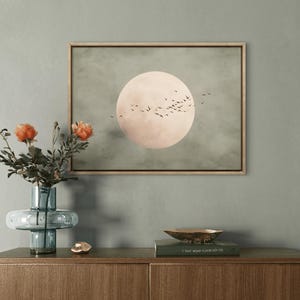 May include: A framed artwork depicts a large, pale pink moon with a flock of birds against a muted green background. A blue glass vase holds orange flowers, and a gold bowl sits on a wooden cabinet, creating a serene, nature-inspired scene.