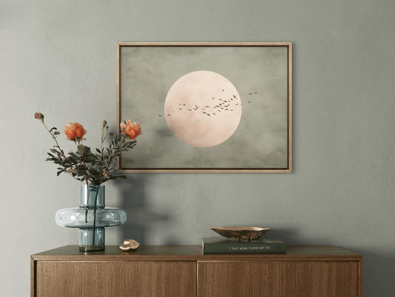 May include: A framed artwork depicts a large, pale pink moon with a flock of birds against a muted green background. A blue glass vase holds orange flowers, and a gold bowl sits on a wooden cabinet, creating a serene, nature-inspired scene.